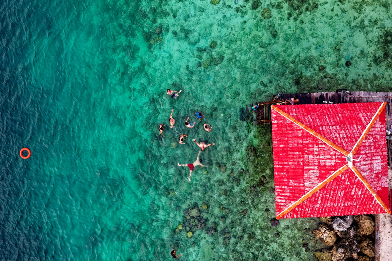 A group enjoying swimming near a vibrant red-roofed hut in turquoise waters.