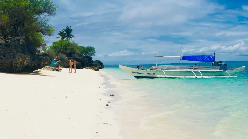 A traditional wooden outrigger banca boat with a blue tarp canopy anchored on the pristine white sand shoreline of Bantayan Island Cebu with clear turquoise water