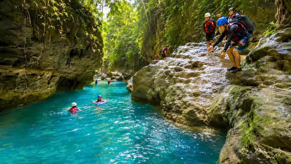 Adventurers wearing helmets and life vests prepare to jump from a rock cliff into bright turquoise river water during a Kawasan Falls canyoneering tour in Badian Cebu