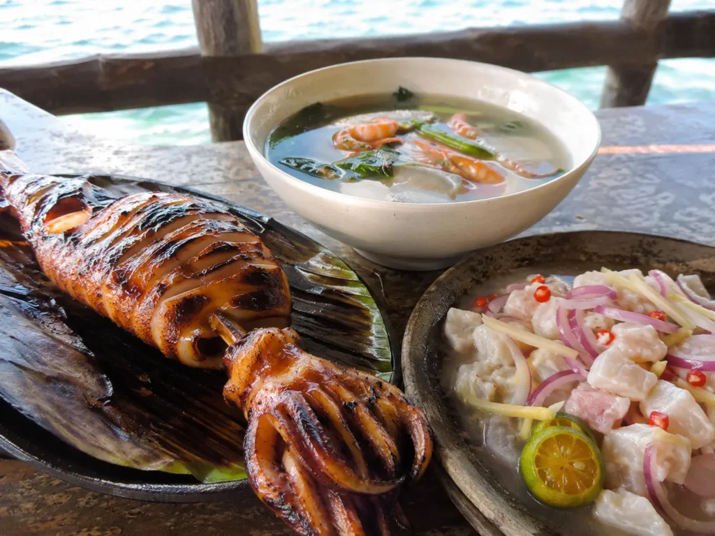 Cebu sutukil seafood spread with grilled squid, seafood soup, and kinilaw on a seaside table.