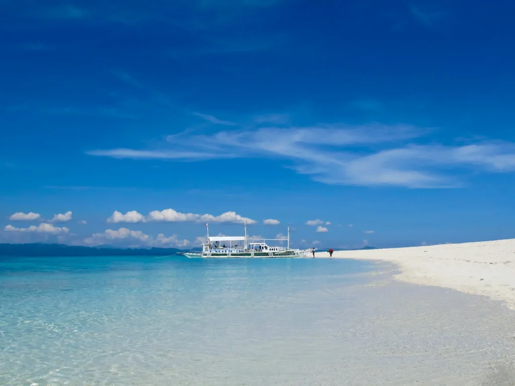 White sand beach on Malapascua Island, Cebu, with clear turquoise water, a small outrigger boat, and a bright blue sky.