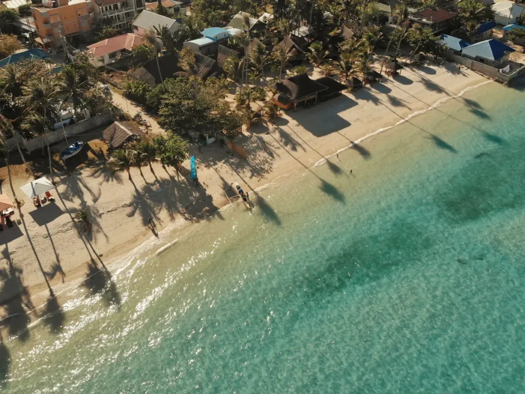 Aerial view of Santa Fe beach in Bantayan Island, Cebu, showing clear turquoise water, white sand, palm trees, and beachfront buildings.